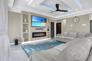 A modern living room with a large flat-screen TV, a fireplace, a ceiling fan, and a beige sofa.