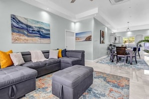 A modern living room with a grey sectional sofa, a dining area with a table and chairs, and a kitchen in the background.