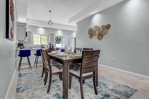 A modern dining area with a wooden table and chairs, a blue rug, and a kitchen in the background.