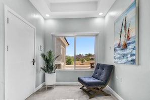 Una habitación con un ventanal grande, una silla, una planta en maceta y un cuadro en la pared.