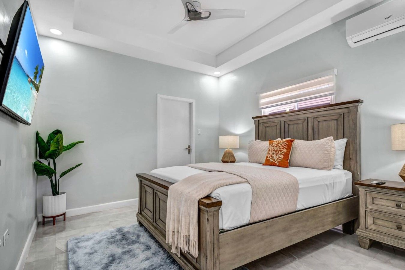 Un dormitorio con cama, mesitas de noche, televisión, planta y ventana con persianas.