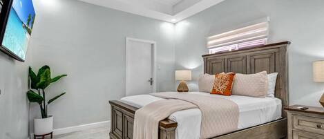Un dormitorio con cama, mesitas de noche, televisión, planta y ventana con persianas.
