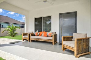 A covered patio area with two wooden chairs and a sofa, a sliding glass door, and a small palm tree.