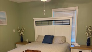 Une chambre à coucher comprenant un lit, des tables de chevet, une commode, un ventilateur de plafond et une fenêtre avec des stores.