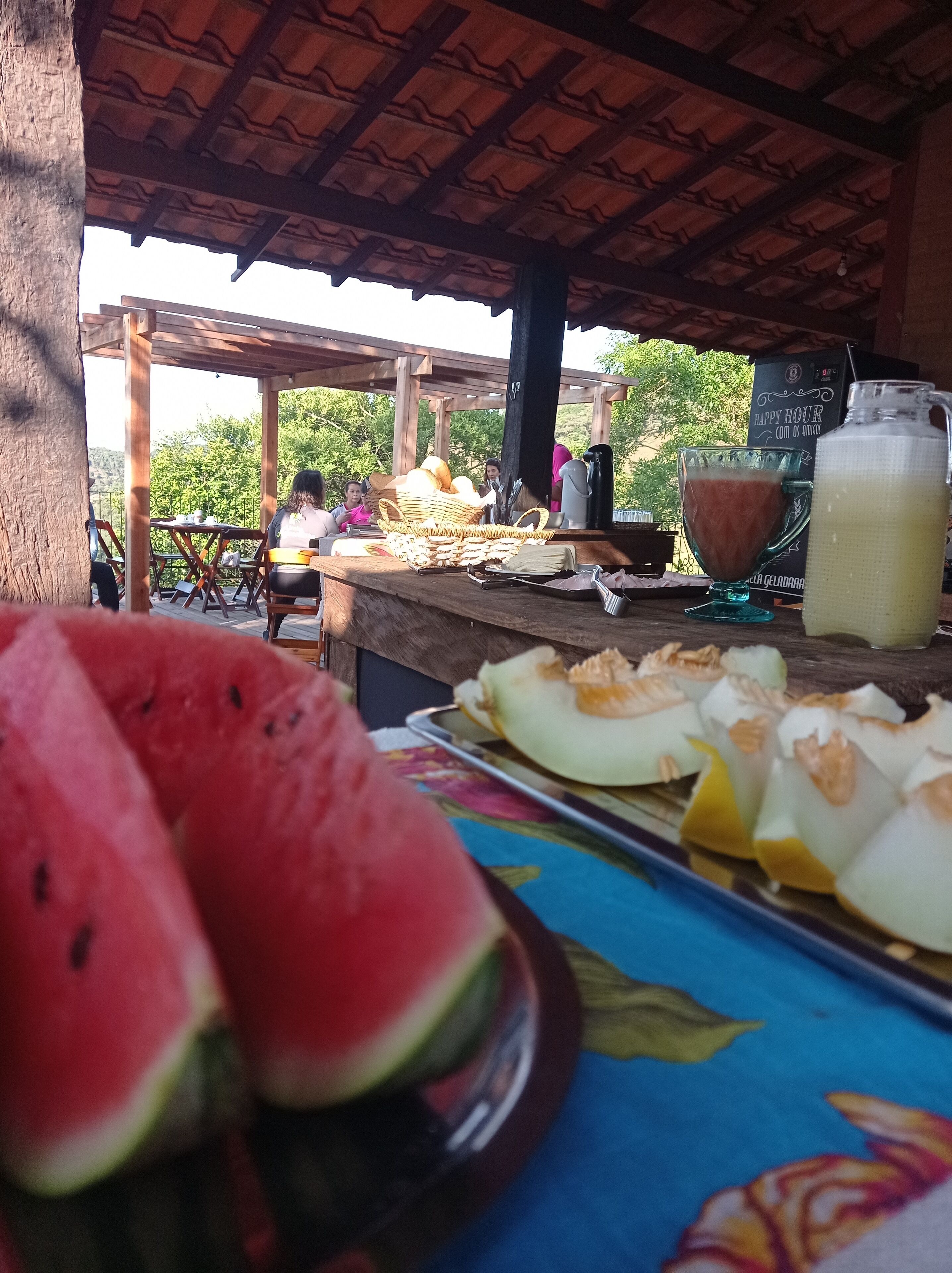 Un área de comedor al aire libre con una mesa de madera, una sandía, fruta cortada y una jarras de jugo.