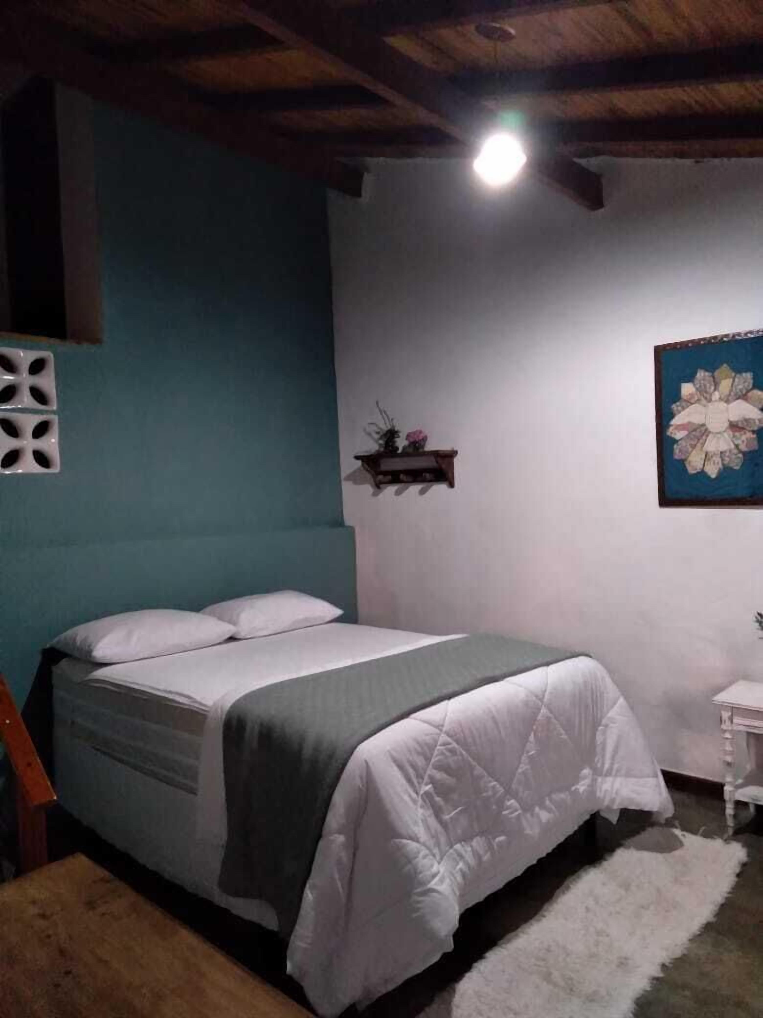 A bedroom with a bed, a small bedside table, a wall-mounted shelf, and a ceiling light.