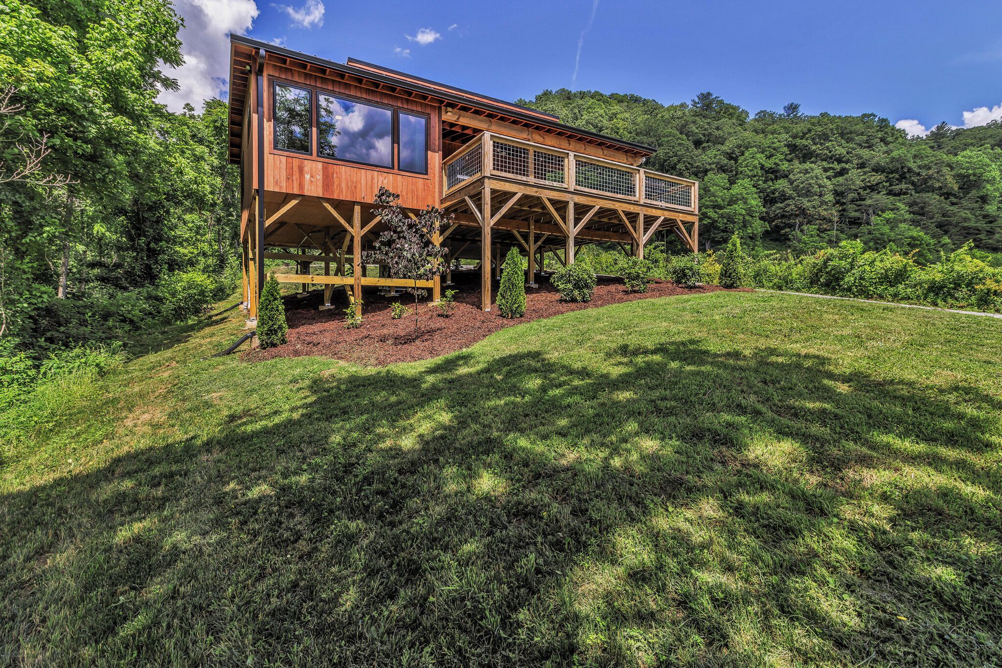 A wooden cabin with a deck, surrounded by lush greenery and a forested hillside.