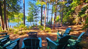 A lakeside campsite with green Adirondack chairs, a fire pit, and a view of the lake surrounded by trees. - Pine Camp*Seaplane/Boat Dock*Lake/Mountain View*Kayaks (Greenville)