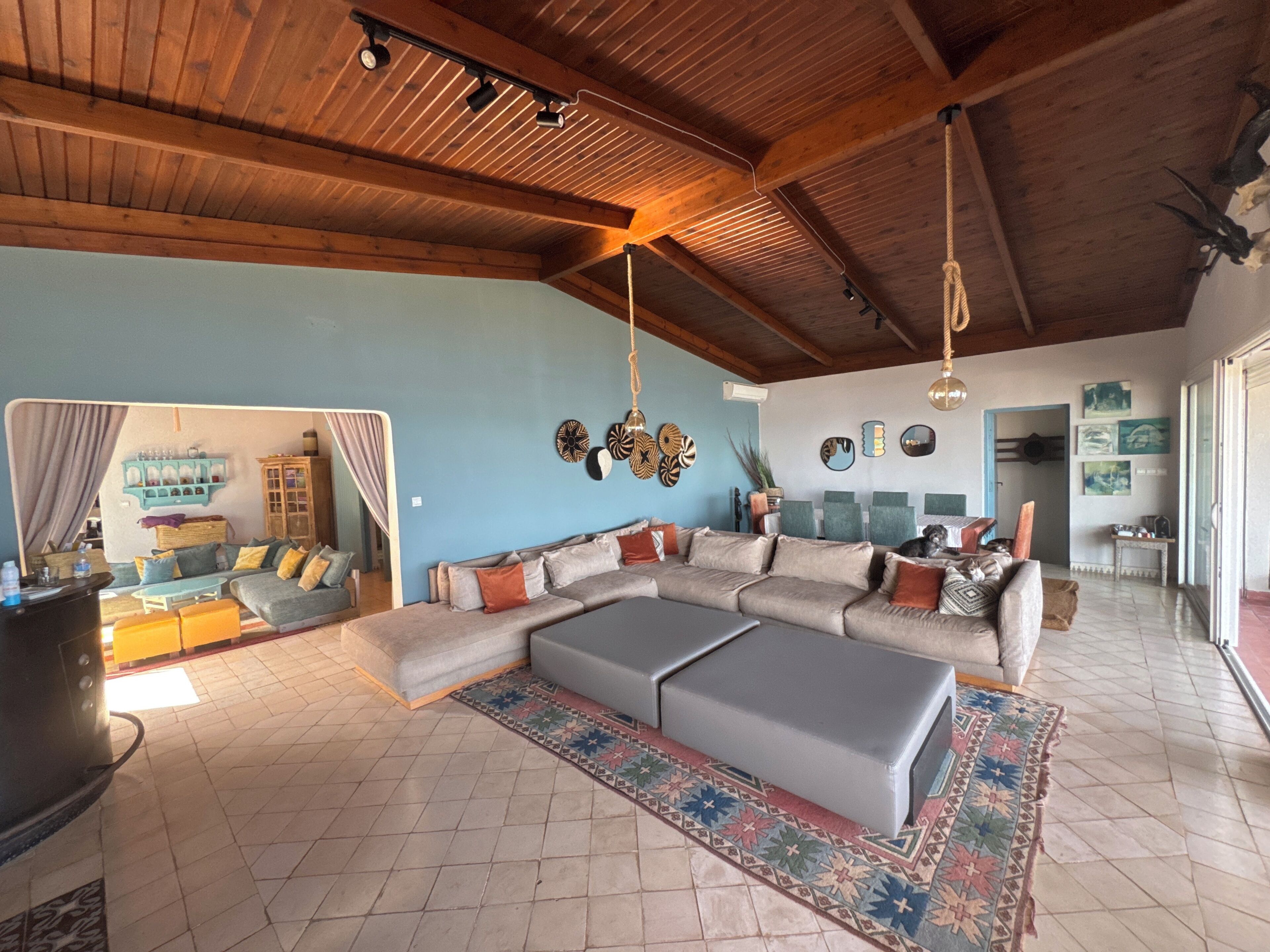 Un vaste espace de vie comprenant un grand canapé d’angle, une table basse et un tapis à motifs. La pièce est agrémentée de poutres apparentes en bois au plafond, d’un mur d’accent bleu et de divers éléments décoratifs.