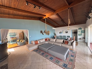 A spacious living area with a large sectional sofa, a coffee table, and a patterned rug. The room features wooden ceiling beams, a blue accent wall, and various decorative items.