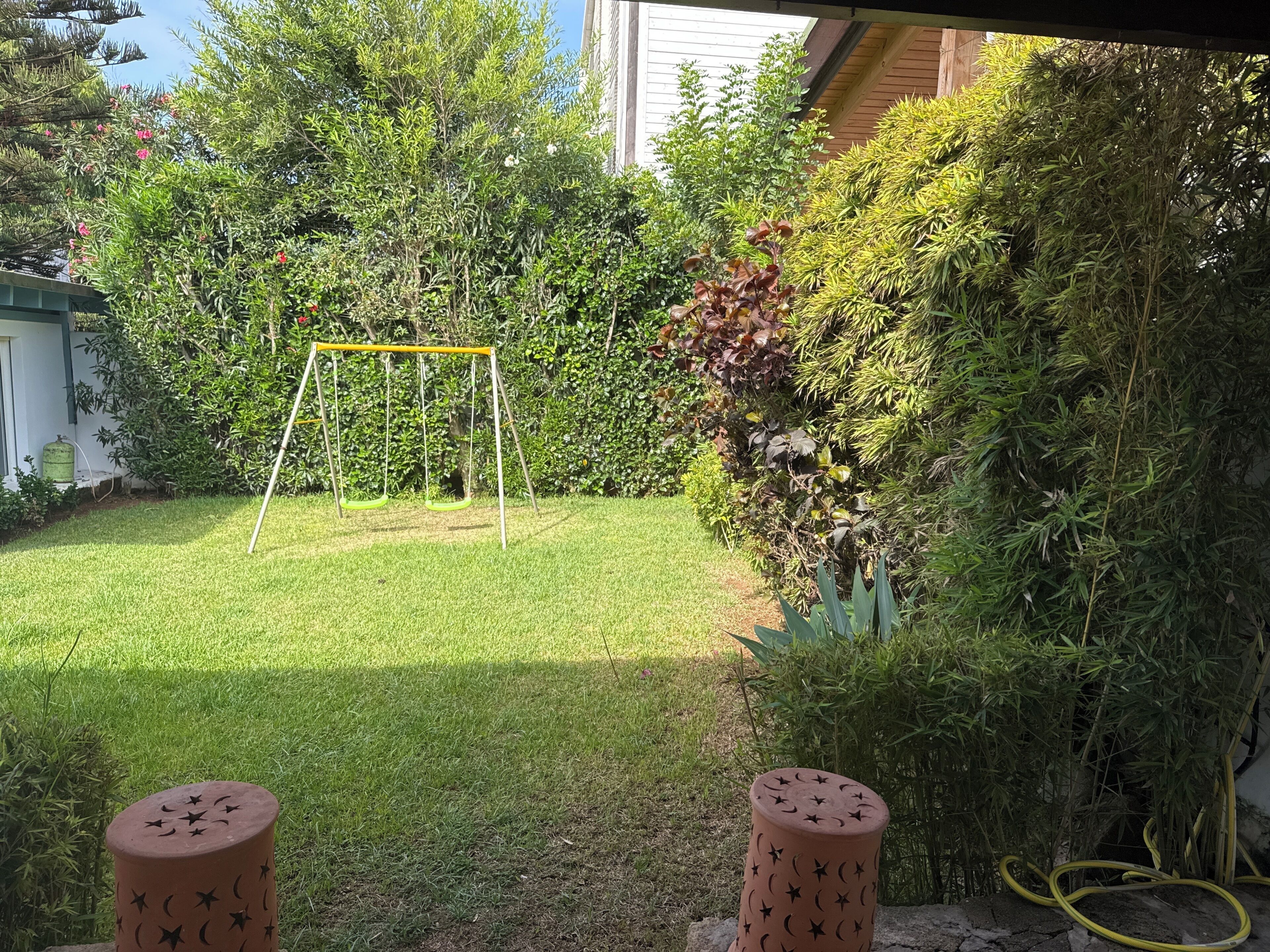 Un jardin à l’arrière de la maison, avec une balançoire, un tuyau d’arrosage et un potager où poussent différentes plantes.
