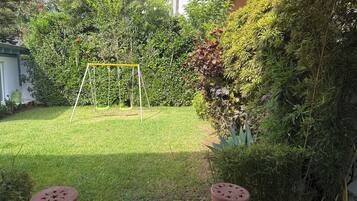 Un jardin à l’arrière de la maison, avec une balançoire, un tuyau d’arrosage et un potager où poussent différentes plantes.