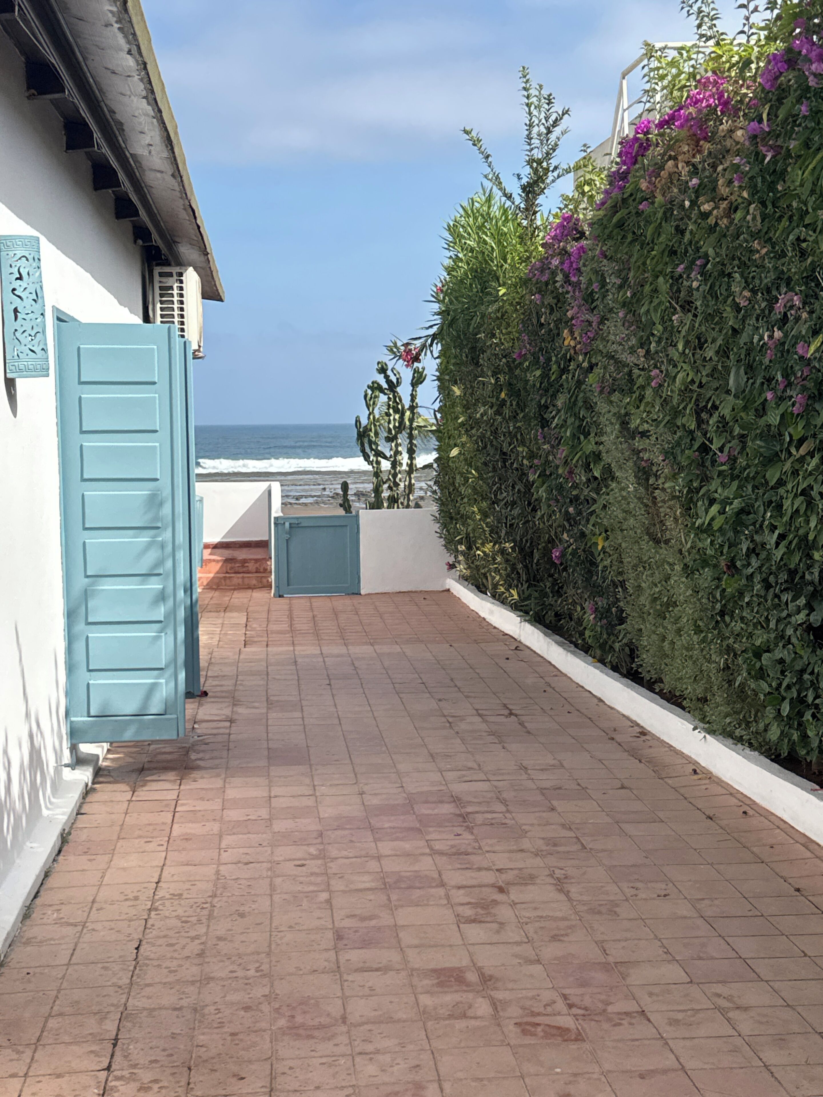 Un chemin pavé mène à un bâtiment doté d’une porte bleue, d’un mur blanc et offrant une vue sur l’océan.