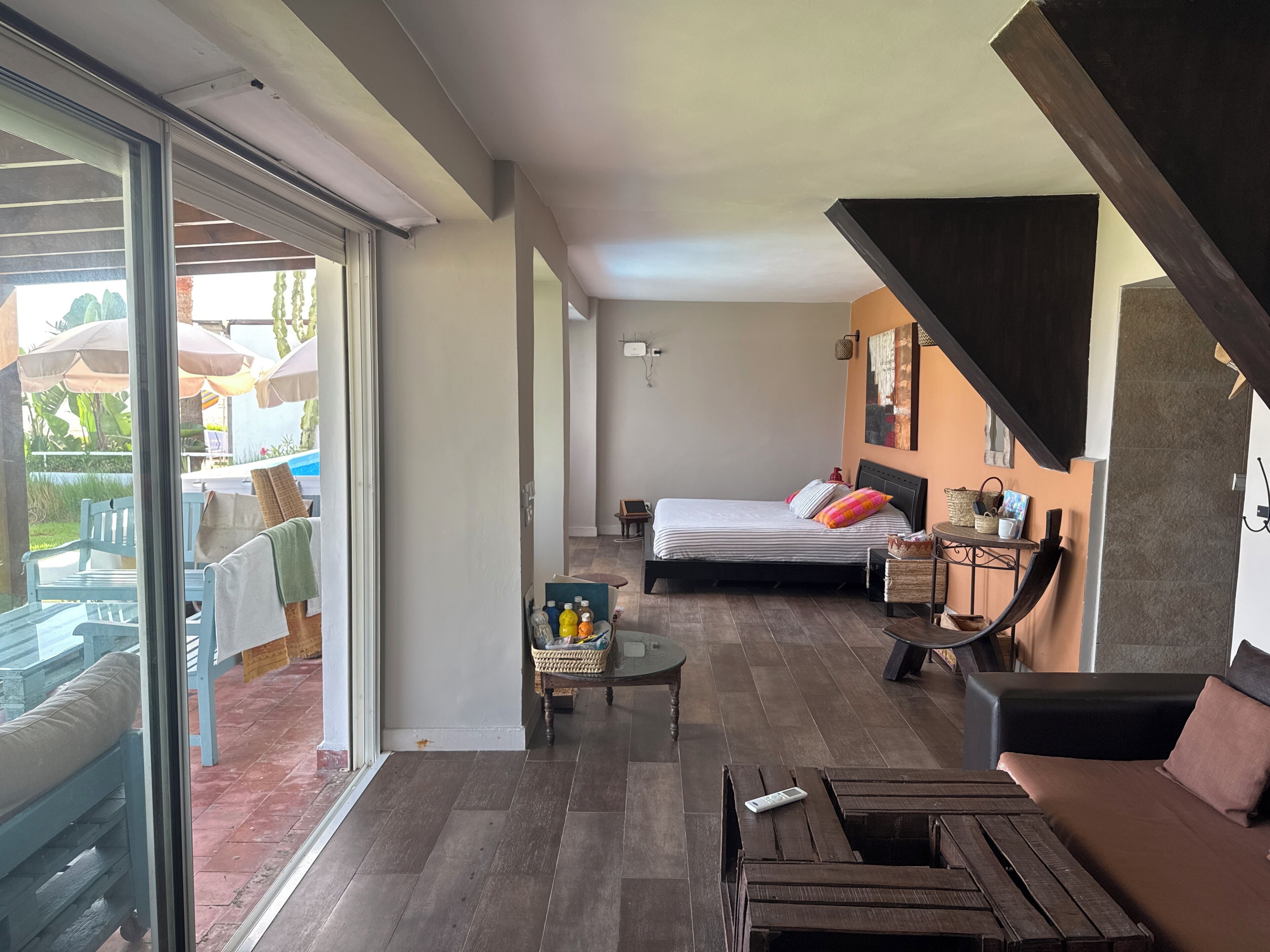 A modern bedroom with a large bed, a small table, and a sofa. There is a sliding glass door leading to an outdoor area with umbrellas.