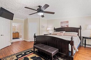 A bedroom with a bed, a bench, a ceiling fan, a TV, and a bathroom visible through an open door.
