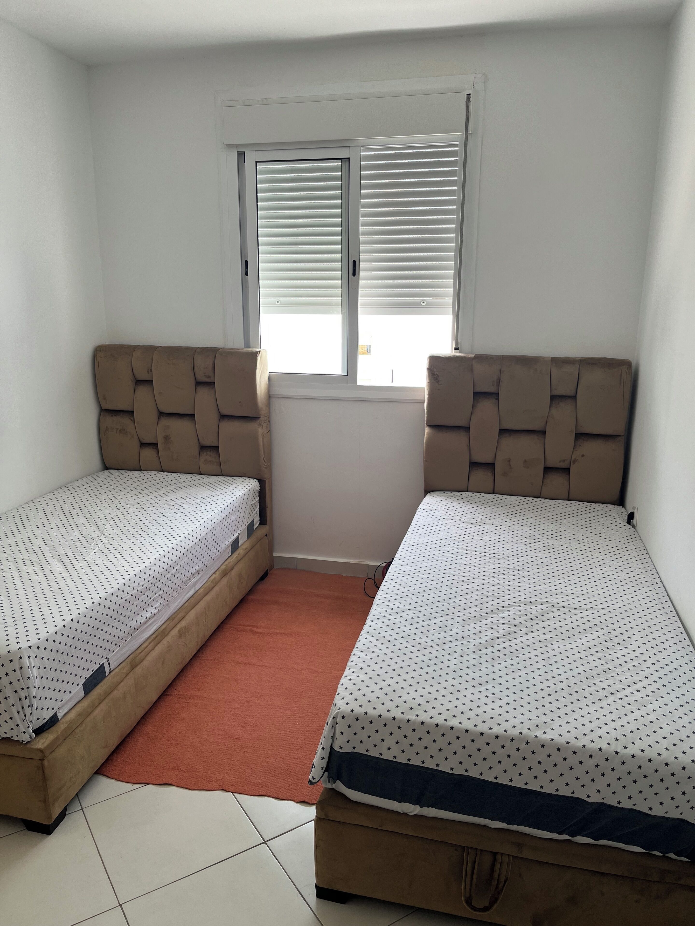Ein kleiner, sauberer Raum mit zwei Einzelbetten, einem Fenster mit Jalousien und einem roten Teppich.