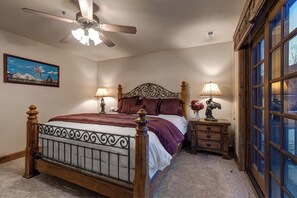 A bedroom with a bed, a ceiling fan, a nightstand with a lamp, a framed picture on the wall, and a window with a view.