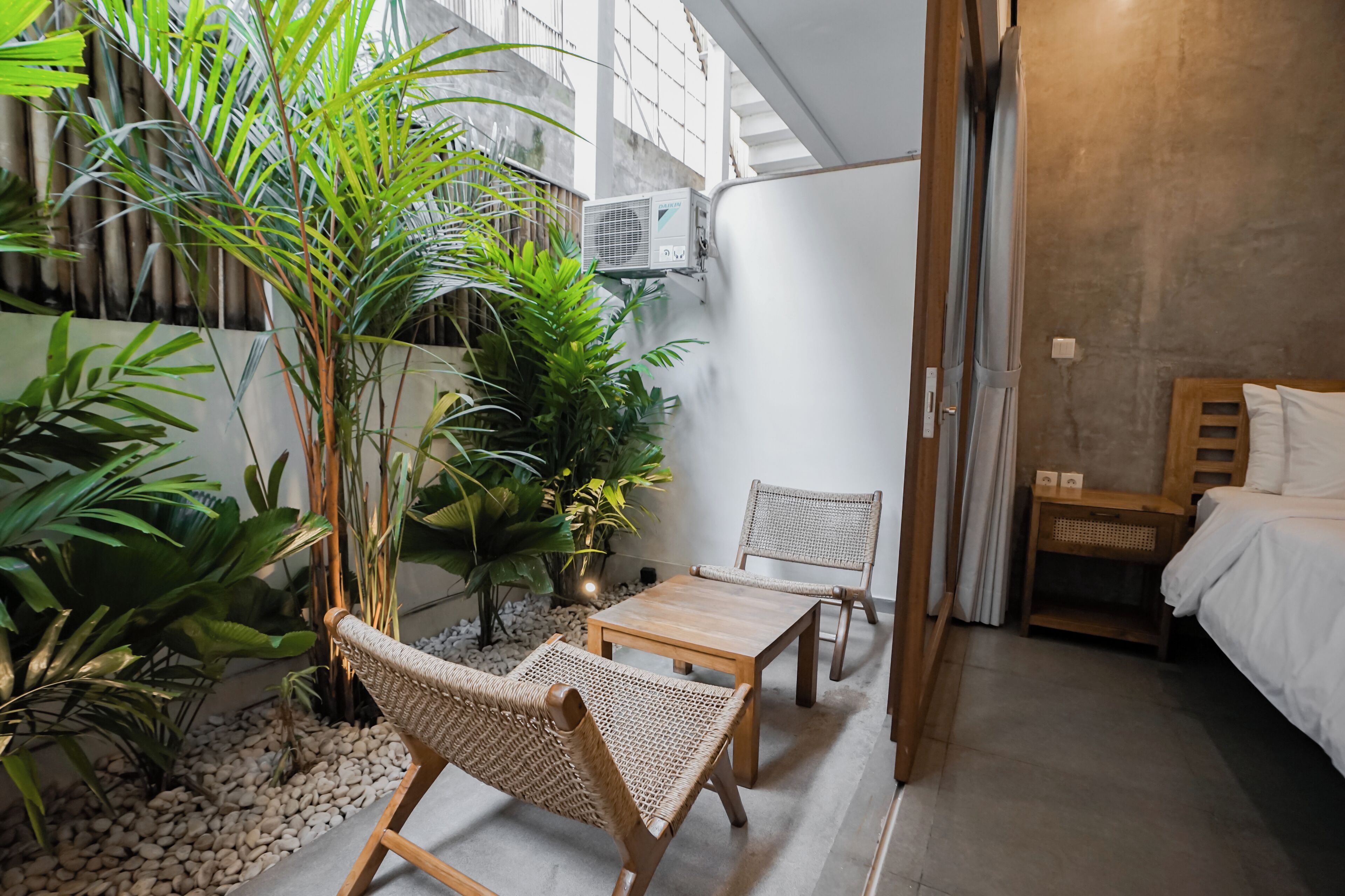 A room with a bed, a small table, two wicker chairs, and a potted plant.