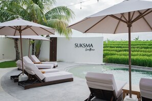A poolside area with lounge chairs and umbrellas, a sign reading "SUKSMA", and a lush green field in the background. - Suksma Guesthouse (Tumbak Bayuh)
