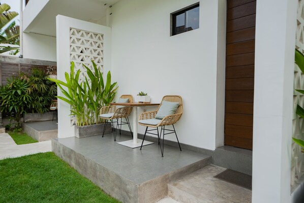 A patio with a table and chairs, a potted plant, and a white wall with a window.