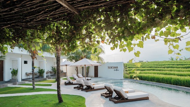 A poolside area with lounge chairs and umbrellas, a well-maintained lawn, and a building with 'SUKISMA' signage. - Suksma Guesthouse (Tumbak Bayuh)