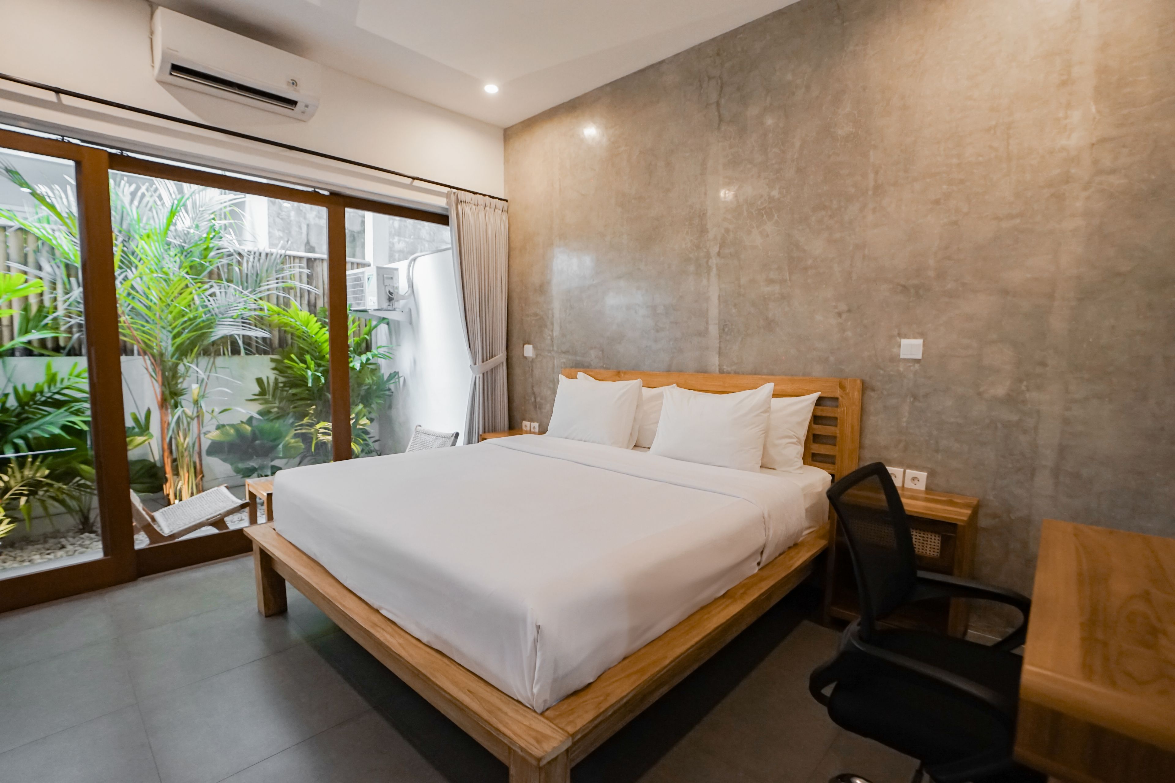 Une chambre avec un grand lit, une table de chevet en bois, un bureau et une chaise. Il y a une fenêtre avec des rideaux et une vue sur la verdure à l’extérieur.