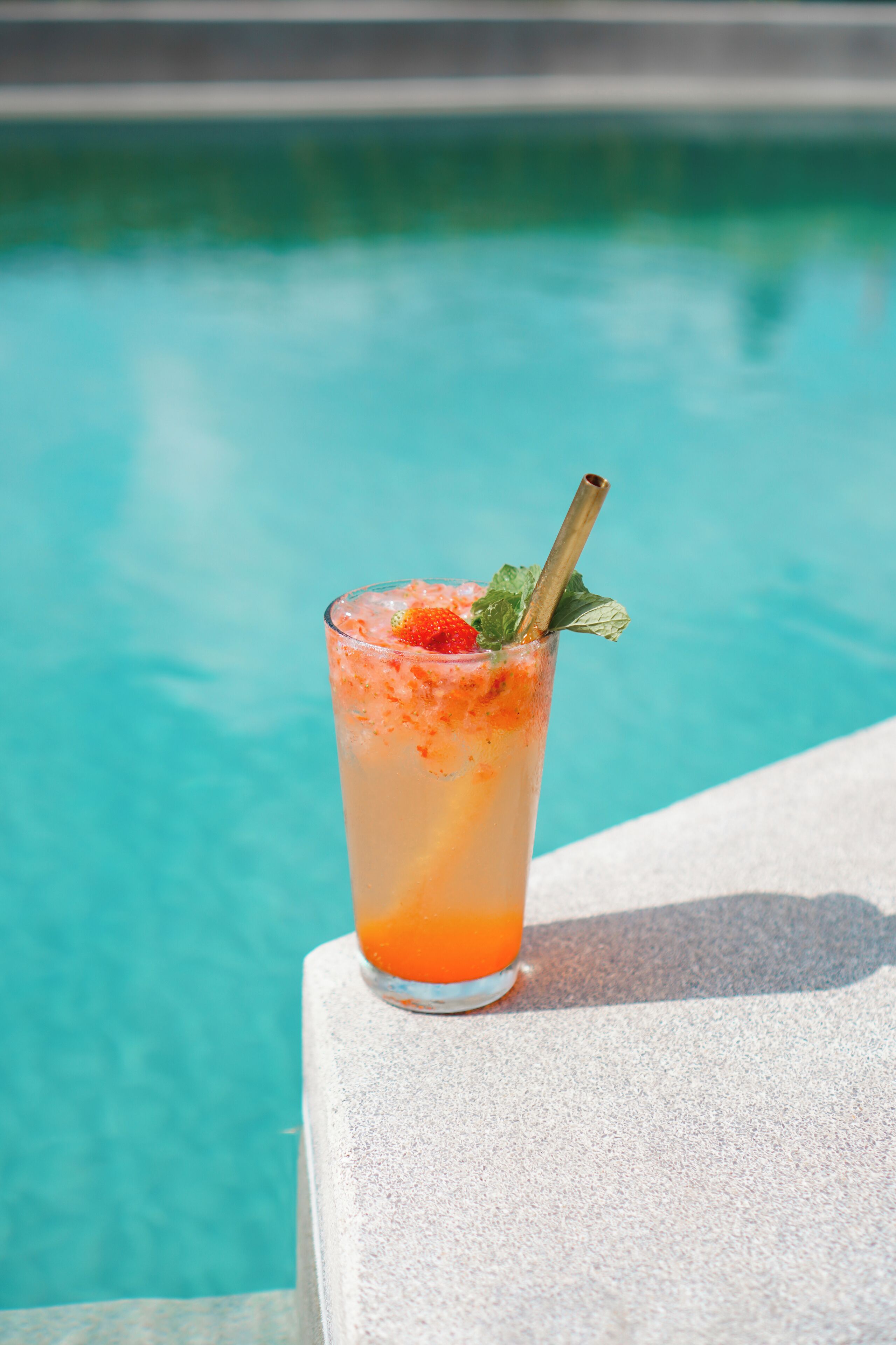 A tall glass with a straw, garnished with a strawberry and mint, placed on a stone ledge by a pool.