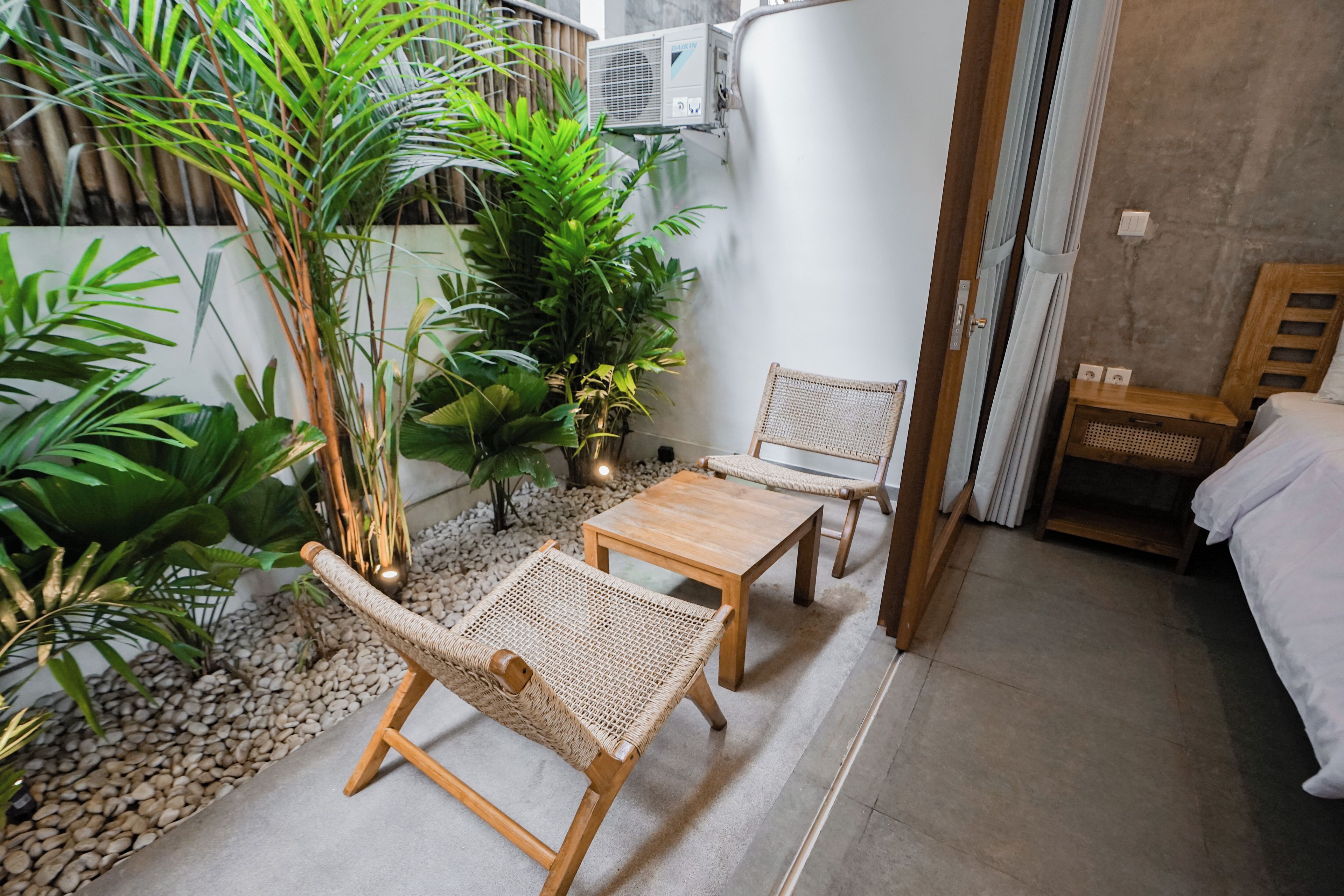 Een knus kamertje met een bed, een houten tafel, twee rieten stoelen en diverse kamerplanten.