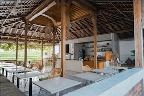 Un área de comedor al aire libre con mesas y sillas, una cocina al fondo y vista a un campo verde.