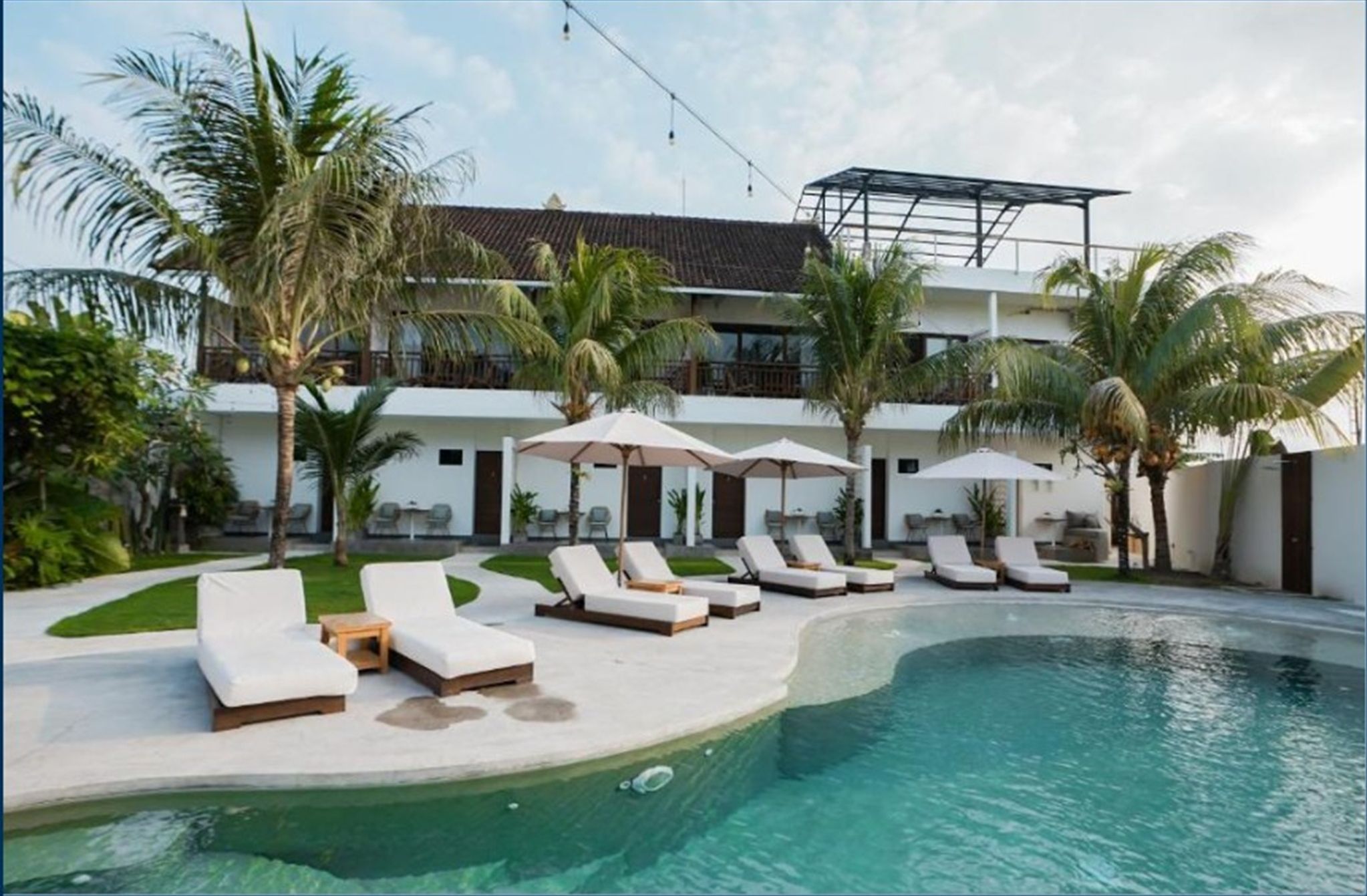 A pool area with sun loungers and umbrellas, surrounded by palm trees and a building with a balcony.
