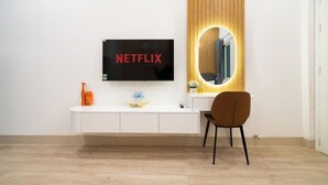 Una habitación de hotel moderna con una mesa consola blanca, un televisor de pantalla plana mostrando 'NETFLIX', un tocador de madera con espejo y una silla marrón.