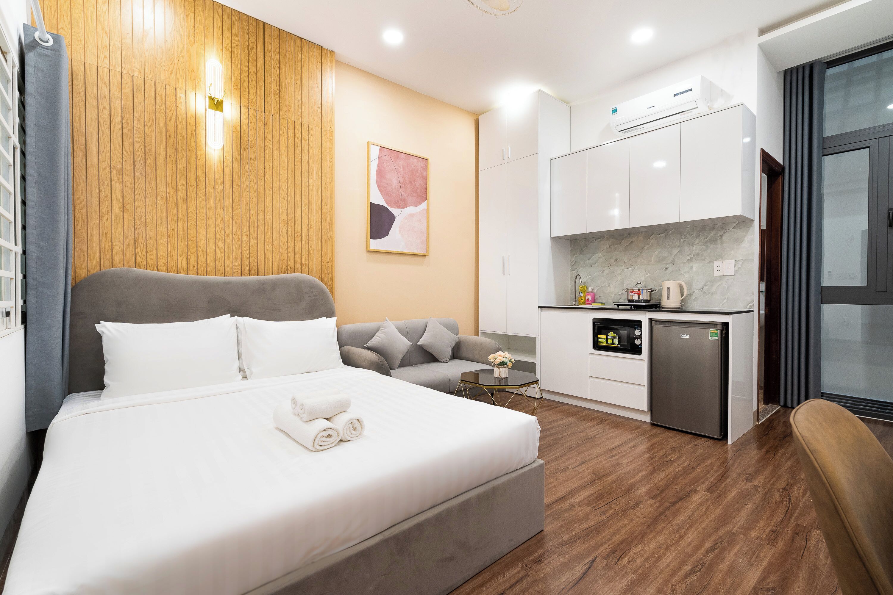 A modern hotel room with a large bed, a sofa, a small kitchen area, and a dining table.