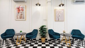 Un área de recepción con piso a cuadros, sillas color aguamarina, una pequeña mesa con una planta y obras de arte enmarcadas en las paredes.