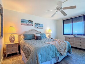 A bedroom with a large bed, two bedside tables with lamps, a ceiling fan, and a view of the ocean through the window.