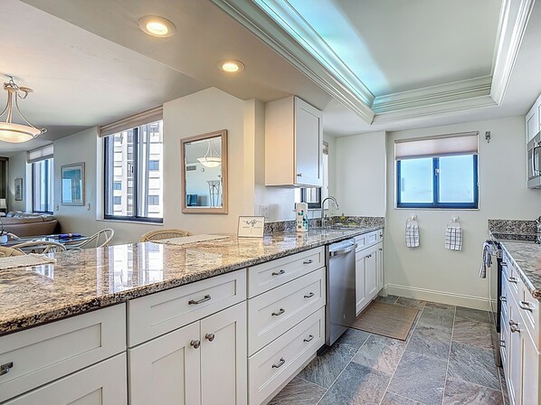 A modern kitchen with granite countertops, white cabinetry, and a large island. There is a dining area with a table and chairs, and a view of the city through large windows.