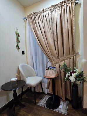 Ein Zimmer mit einem beigen Vorhang, einem weißen Stuhl, einem kleinen runden Tisch mit schwarzem Gestell, einem kleinen Holztisch mit einem Korb und einer Vase mit Blumen.