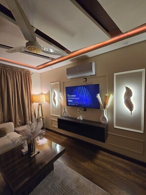 A modern living room with a flat-screen TV, decorative wall art, a ceiling fan, and a mounted air conditioner. - Shahzaib's Home 1BHK Modern Apartment at City Center (Lahore)