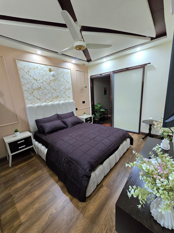 Ein modernes Schlafzimmer mit einem großen Bett, einem Deckenventilator, einem Nachttisch, einem Schreibtisch mit Blumen und einer Tür zu einem weiteren Raum.