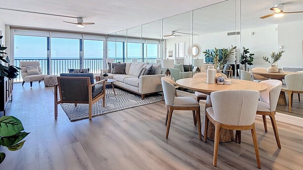 A spacious living area with a large window, a dining table, and a sofa. - Beachfront Getaway at Caper Beach Club #813 | Private Chiller/Heated Pool (Fort Myers Beach)