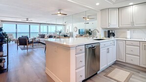 A modern kitchen with white cabinetry, a large island, and a dishwasher. There is a dining area with a sofa and a coffee table. - Beachfront Getaway at Caper Beach Club #813 | Private Chiller/Heated Pool (Fort Myers Beach)