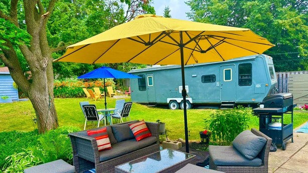 Un patio al aire libre con mobiliario de mimbre, un paraguas amarillo y una autocaravana azul en el fondo.