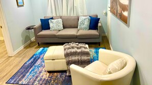 Una sala de estar con un sofá color beige, dos almohadas azules, un sillón blanco, un tapete estampado y un cuadro en la pared.