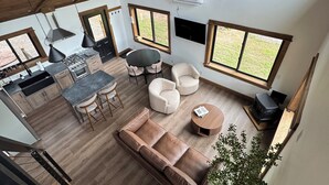 En rummelig opholdsstue med højt til loftet, træbjælker, et køkken med moderne hårde hvidevarer og store vinduer med udsigt til naturen.