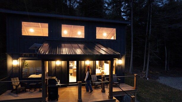 A two-story wooden cabin with a covered deck, outdoor seating, and a visible kitchen area. - Hemlock Hideaway – Luxury Cabin - Romantic & Remote Getaway in Upstate, NY (Otego)