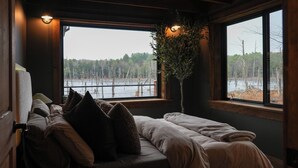 A cozy room with a bed, a window with a view of a forest and a body of water, and a potted plant. - Hemlock Hideaway – Luxury Cabin - Romantic & Remote Getaway in Upstate, NY (Otego)