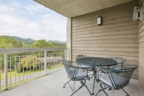 A balcony with a metal table and chairs, a white railing, and a view of a green landscape with hills.