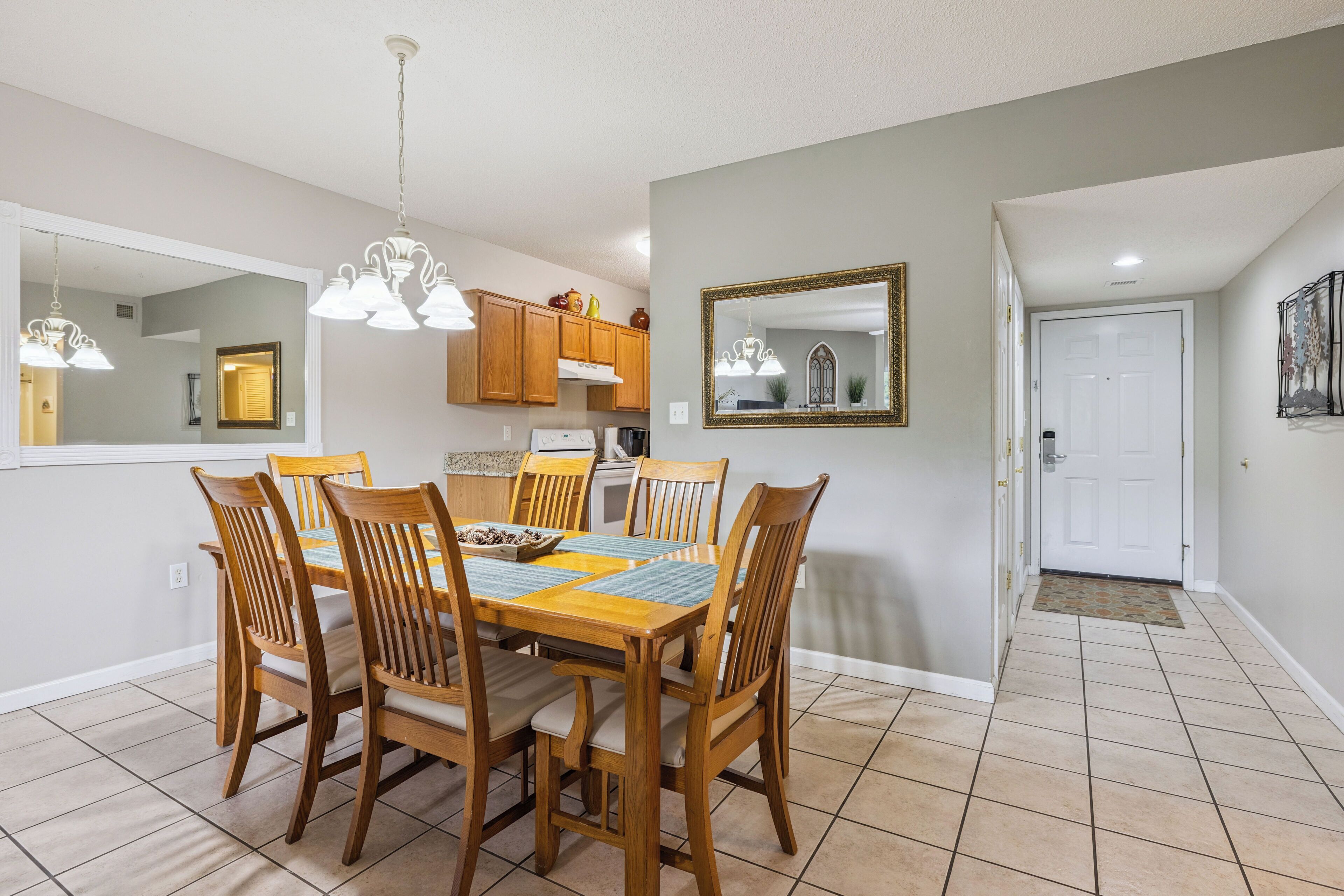 A dining area with a wooden table and chairs, a chandelier, and a kitchen in the background.