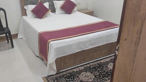 Un dormitorio con cama, almohadas, una silla, una mesita de noche y un cubo rojo.