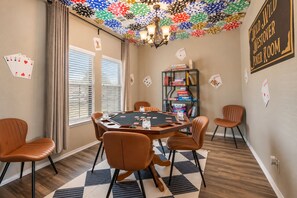 A poker room with a black felt table, brown leather chairs, a bookshelf with books, and a ceiling decorated with poker chips.