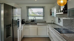 Een moderne keuken met witte kasten, een roestvrijstalen koelkast, een granieten aanrechtblad en een groot raam met zonwering.